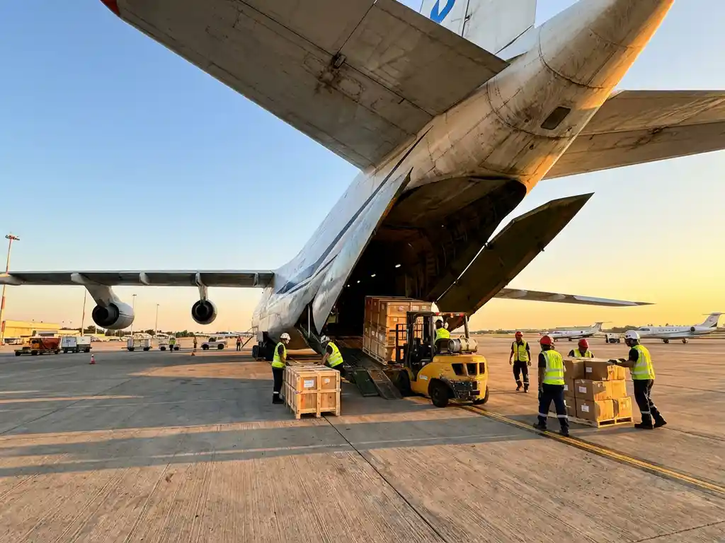 Vrachttoestel met open laaddeuren terwijl grondpersoneel dozen en pallets lost op vliegveldplatform met vorkheftruck